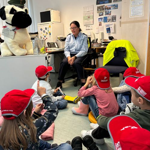 Kinder mit roten Mützen sitzen auf dem Boden in einem Büro, hören einer uniformierten Frau vor ihrem Schreibtisch aufmerksam zu.