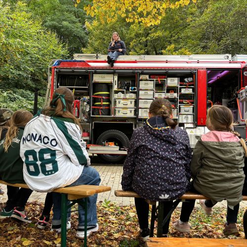 Kindergruppe sitzt auf Bänken vor einem offenen Feuerwehrfahrzeug, auf dem eine Frau mit Mikrofon spricht, umgeben von Wald.