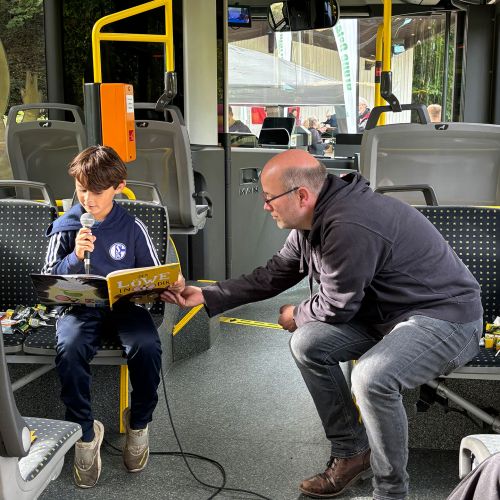 Junge liest mit Mikrofon in einem Bus vor, ein Mann hält das Heft offen.