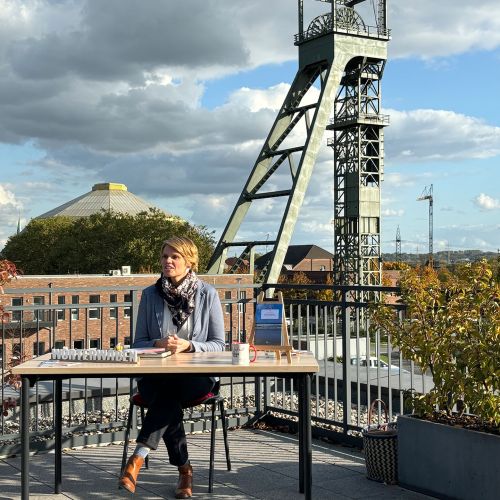 Frau sitzt an einem Tisch auf einer Dachterrasse, im Hintergrund der Förderturm der Zeche Osterfeld.